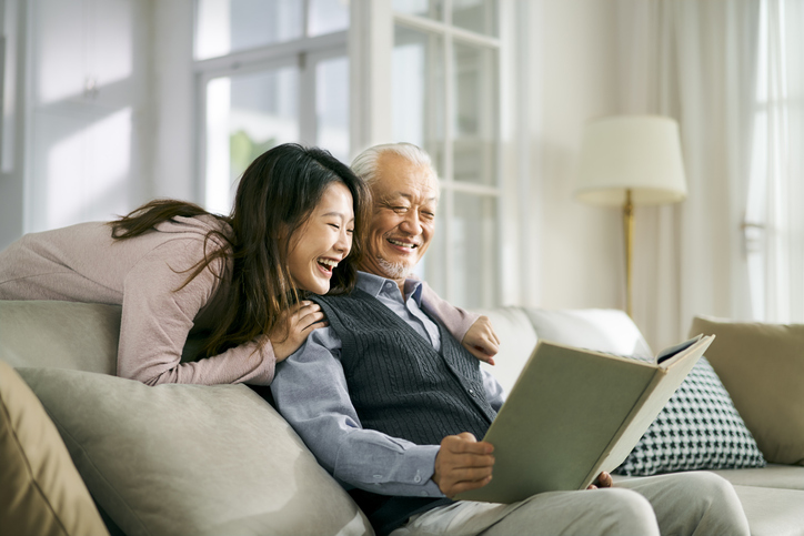 a senior father and his adult daughter reading a book together on a couch